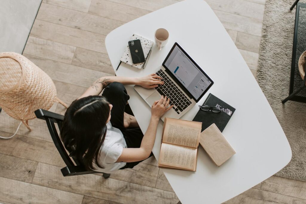 Vue aérienne d'une femme utilisant un ordinateur portable à un bureau à la maison, entourée de livres, d'un téléphone et d'une tasse.